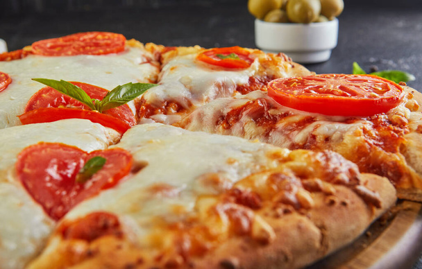 Włoska pizza i składniki do gotowania na czarnym betonowym tle. Pomidory, oliwki, bazylia i przyprawy. Pokrojony trójkąt pizzy. Widok z boku. Zamknij się. - Zdjęcie, obraz