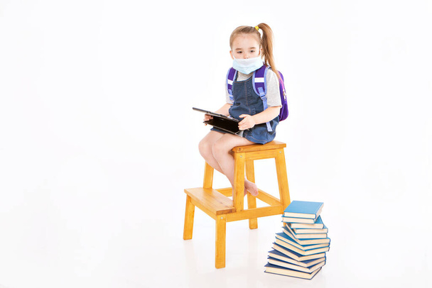 first-grader in medical mask learns to read. little girl on home distance learning. child in medical mask works at laptop and books. doing homework. isolated on white background. knowledge day - Fotó, kép
