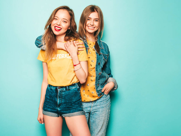 Two young beautiful smiling hipster girls in trendy summer yellow T-shirts and jeans jacket. Sexy carefree women posing near blue wall. Trendy and positive models having fun - Фото, изображение
