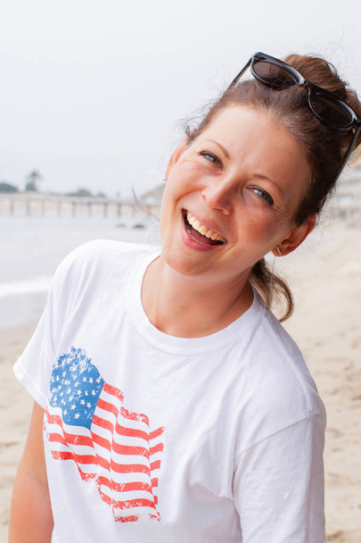 Bella giovane donna in t-shirt bianca con la bandiera americana che cammina lungo la spiaggia di sabbia in una giornata di sole. Concetto di libertà e stile di vita felice - Foto, immagini