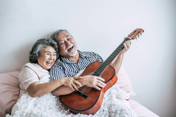 Το ηλικιωμένο ζευγάρι χαλαρώνει παίζοντας ακουστική κιθάρα στο κρεβάτι - Φωτογραφία, εικόνα
