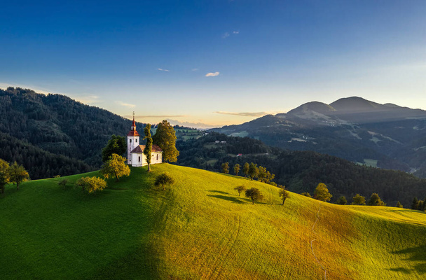 Skofja Loka, Slowenien - Luftaufnahme der wunderschönen Kirche Sveti Tomaz (Sankt Thomas) mit einem warmen Sommersonnenuntergang, klarem blauen Himmel und den Julischen Alpen im Hintergrund - Foto, Bild