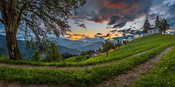 Skofja Loka, Slovenya - Sloven Alpleri 'nin büyüleyici tepelik kilisesi Sveti Tomaz (Saint Thomas), renkli gökyüzü ve yazın arka planda Julian Alps ile güzel panoramik kırsal manzarası - Fotoğraf, Görsel