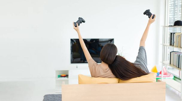 Két fiatal ázsiai nő kezében játék joysticks és versenyeznek boldogan, miközben ül a kanapén. Versenyképes barátok videojátékkal otthon. - Fotó, kép