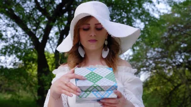 Крупным планом женщина в белой шляпе открывает коробку с подарком, удивляется и улыбается
 - Кадры, видео