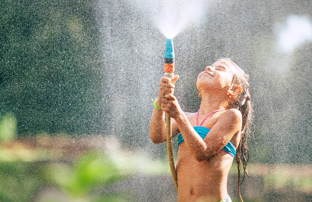 Cute little girl pours herself from the hose, makes a rain. Hot summer day pleasure - Фото, изображение