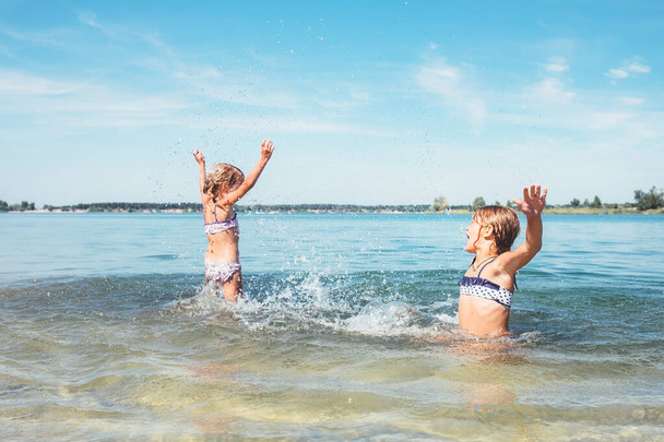 Two little sister girls fooling around in the calm sea waves splashing water to each other. Family vacation concept image. - Zdjęcie, obraz