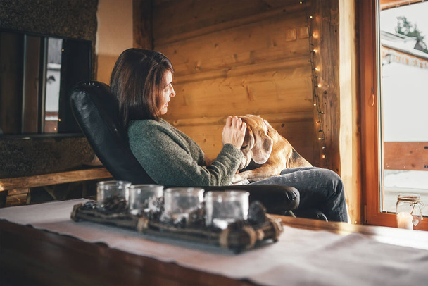 Tender scene with woman and her beagle dog  in comfortable chair opposite big window in cozy country home. Countryside vacation with pets concept image.  - Zdjęcie, obraz