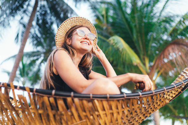 Happy smiling young woman in straw hat in sunglasses sitting in hammock with palm trees background  - Fotó, kép