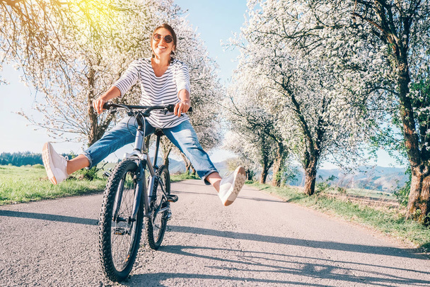 Happy smiling woman cheerfully spreads legs on bicycle on the country road under blossom trees. Spring is comming concept image. - Φωτογραφία, εικόνα