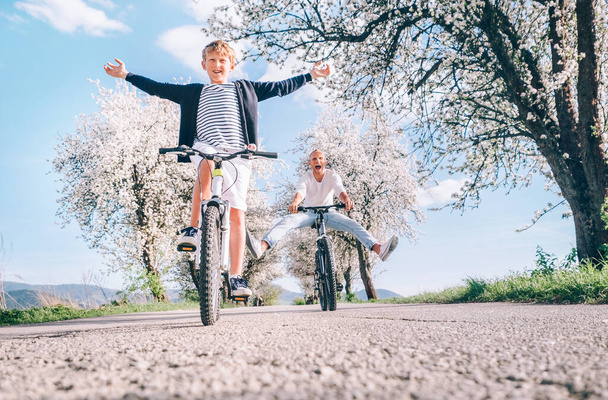 Father and son having fun spreading wide legs and screaming when riding bicycles on country road under blossom trees. Healthy sporty lifestyle concept image. - Photo, image