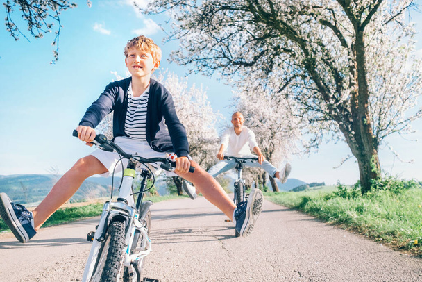 Father and son having fun spreading wide legs when riding bicycles on country road under blossom trees. Healthy sporty lifestyle concept image. - Photo, image