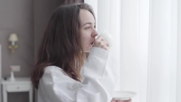 Side view of brunette Caucasian woman drinking coffee in the morning in hotel room. Close-up portrait of positive young girl enjoying vacations at tourist resort. Happiness, tourism, lifestyle, joy. - Imágenes, Vídeo