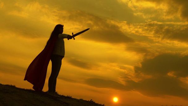 girl plays roman lenin in bright rays of sun against sky. free woman knight. super woman with a sword in his hand and in a red cloak stands on mountain in sunset light. free woman playing superhero. - Photo, Image