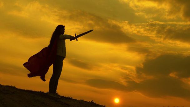 girl plays roman lenin in bright rays of sun against sky. free woman knight. super woman with a sword in his hand and in a red cloak stands on mountain in sunset light. free woman playing superhero. - Photo, Image