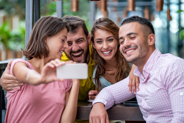 Ryhmä nuoria ystäviä ottaa selfien kahvilassa. He hymyilevät ja pitävät hauskaa yhdessä. Ystävät, vapaa-ajan ja elämäntavan käsite. - Valokuva, kuva