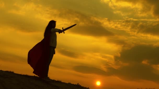 dívka hraje římské lenin v jasných paprscích slunce proti obloze. svobodný rytíř. super žena s mečem v ruce a v červeném plášti stojí v zapadajícím slunci na hoře. volná žena hraje superhrdinu. - Záběry, video