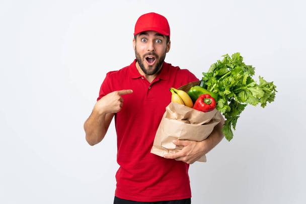 Mladý muž s vousy drží pytel plný zeleniny izolované na bílém pozadí s překvapivým výrazem obličeje - Fotografie, Obrázek