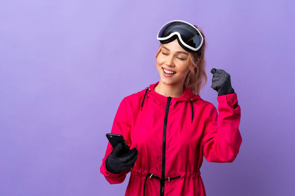 skiër tiener meisje met snowboarden bril over geïsoleerde paarse achtergrond met telefoon in overwinningspositie - Foto, afbeelding