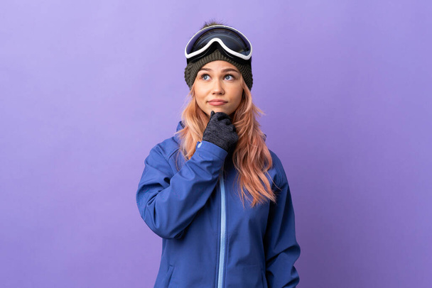 Skier έφηβος κορίτσι με γυαλιά snowboarding πάνω από απομονωμένο μωβ φόντο έχουν αμφιβολίες και τη σκέψη - Φωτογραφία, εικόνα
