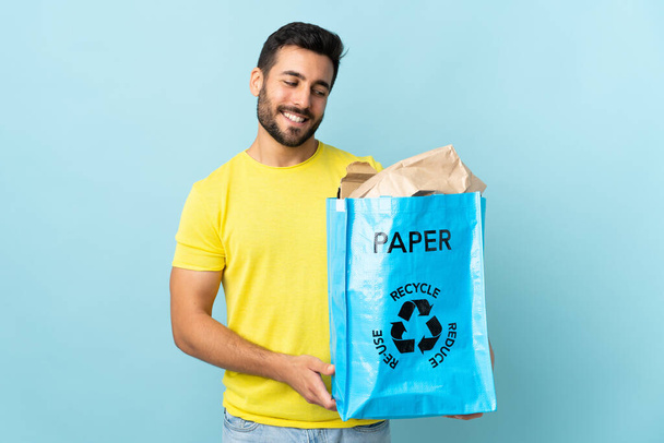 Joven hombre caucásico sosteniendo una bolsa de reciclaje aislado sobre fondo azul con expresión feliz - Foto, imagen