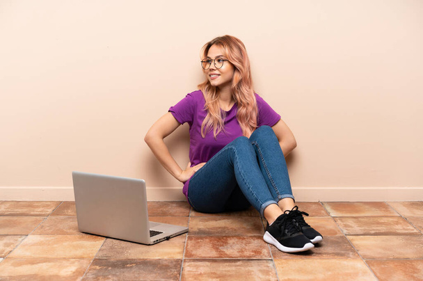 Adolescente donna con un computer portatile seduto sul pavimento al chiuso in posa con le braccia all'anca e sorridente - Foto, immagini