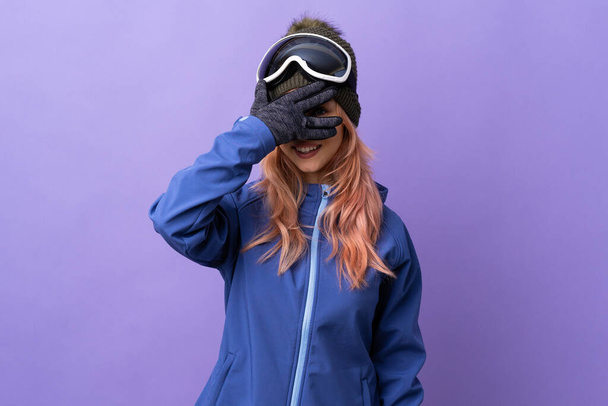 Esquiador adolescente con gafas de snowboard sobre fondo púrpura aislado cubriendo los ojos por las manos y sonriendo
 - Foto, Imagen