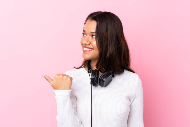 Young woman with earphones over isolated pink wall pointing to the side to present a product - Photo, image