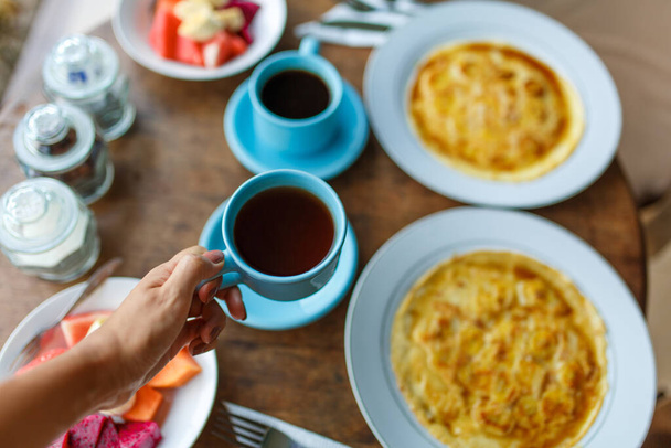 Plates with banana pancakes, tropical fruits and two cups of coffee on wooden table. - Fotoğraf, Görsel
