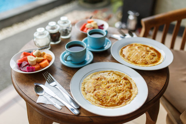 Plates with banana pancakes, tropical fruits and two cups of coffee on wooden table. - Foto, immagini