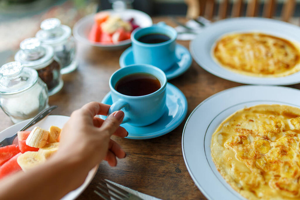 Plates with banana pancakes, tropical fruits and two cups of coffee on wooden table. - Photo, Image