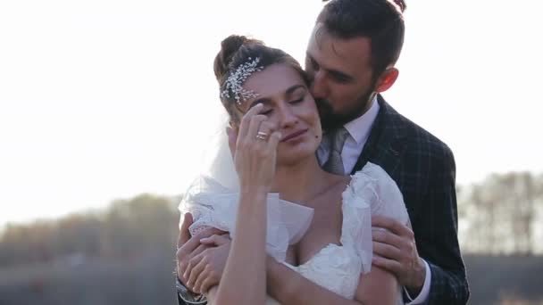 Close-up view of beautiful young wedding couple spending time together in nature on warm sunny autumn day. Happy groom hugs and kisses from behind his beautiful bride in a dress on field background. - Footage, Video