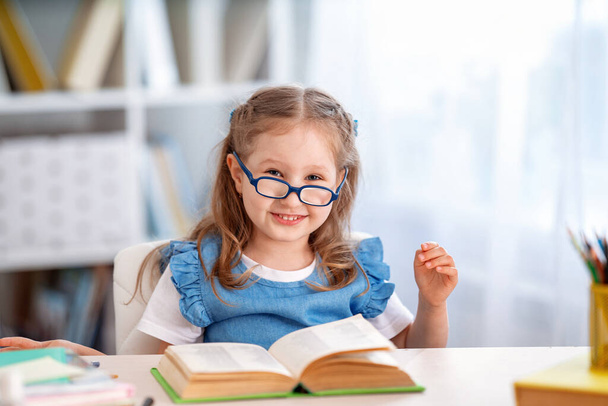 Back to school! A happy smart little girl with glasses is sitting at a table with a book in the room. The child is preparing for school. Do homework. The concept of home schooling. - Φωτογραφία, εικόνα