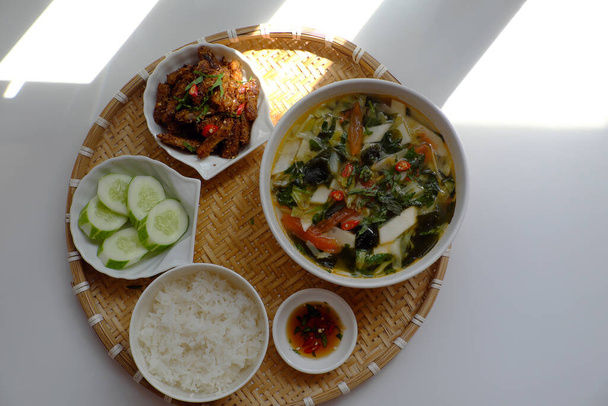 Top view tray of Vietnamese meal for lunch on white background, vegetarian homemade food with seitan fried with citronella, chili, bowl  of vegetable soup and rice dish, simple cuisine from vegetal - Photo, Image