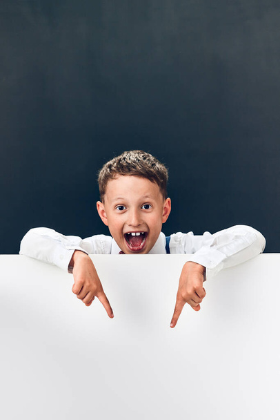 Studio shot of a cheerful Caucasian boy looking out from behind a banner. Happily shouts open mouth and points fingers at an empty space for the text. Advertising and attracting attention - Foto, afbeelding