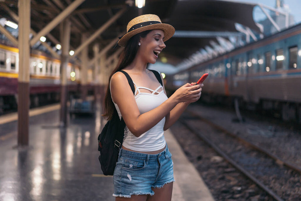 Young brunette woman traveler laughs while playing a mobile phone in train station platform. - Photo, Image