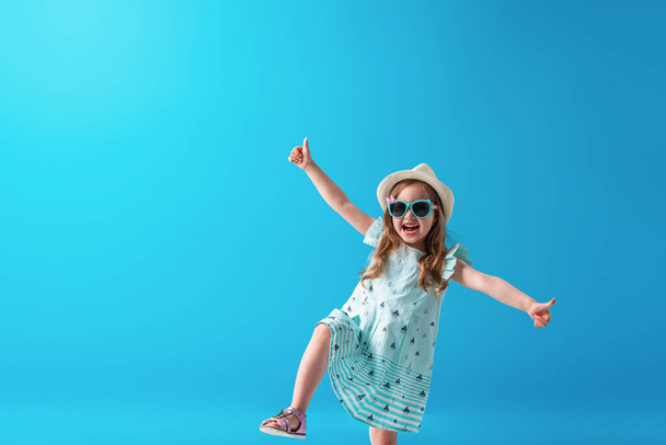 a cute little girl of 4 years old, in a dress, hat and sunglasses, poses, on a blue background. The child will be happy to start the summer holidays and go on a trip - Foto, Imagem