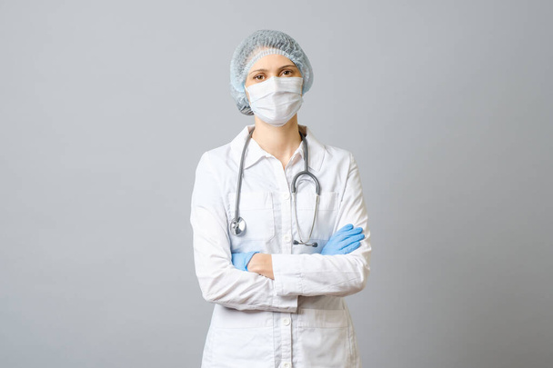 Portrait of young female doctor in protective medical mask on her face and cap on her head. Isolated on gray background - Photo, Image