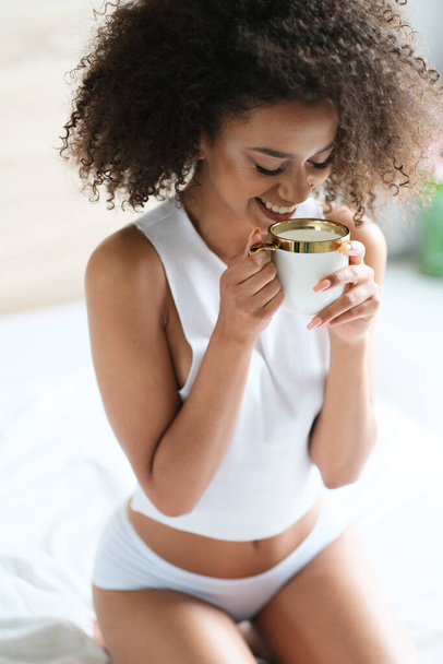 Good morning. Pretty afroamerican model with cup of coffee start a day in a bed. Model in white underwear. - Zdjęcie, obraz