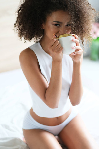 Good morning. Pretty afroamerican model with cup of coffee start a day in a bed. Model in white underwear. - 写真・画像