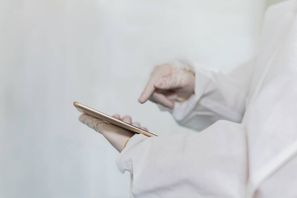 A healthcare worker wearing a protective biohazard suit and gloves using a mobile phone to analyze data on the rapid spread of coronavirus - Zdjęcie, obraz