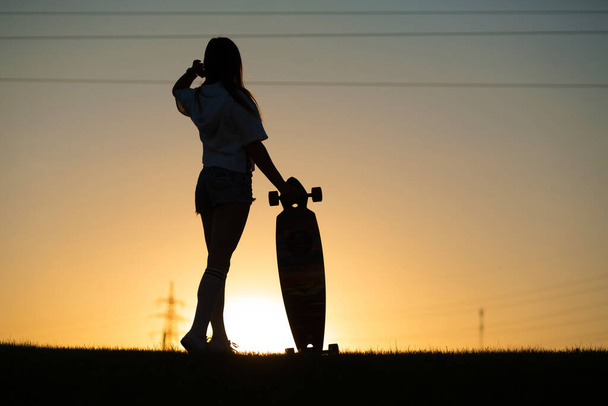 tyttö katsoo auringonlaskua pitäen longboard kädessään. - Valokuva, kuva