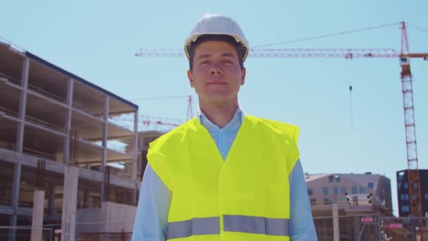 Professional builder standing in front of construction site. Foreman in hardhat helmet and vest. Office building and crane background. Business, real estate and investment concepts. - Materiał filmowy, wideo