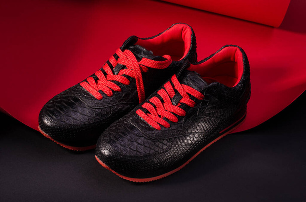 Fashionable snakeskin leather sneakers. Black sneakers with bright red bootlaces against a black and red background and long shadows. - Photo, Image