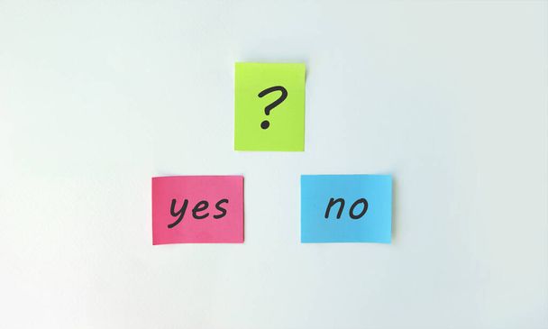 Photo two stickers with the phrase yes or no and with a sticker with a question mark on a white background. Yellow, pink and blue self-adhesive stickers. - Foto, afbeelding