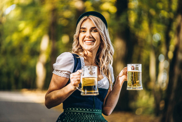 Pretty happy blonde in dirndl, traditional festival dress, holding two mugs of beer outdoors in the forest with blurred background. Oktoberfest, St Patricks day, international beer day concept. - 写真・画像