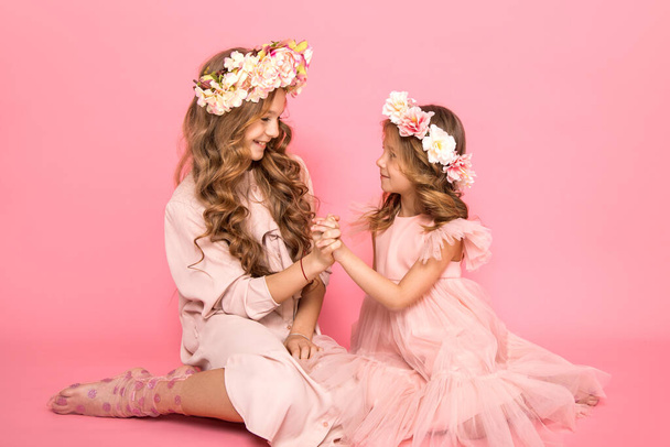 Photo of two beautiful girls are sitting on pink background looking at each other and holding hands. The relationship between an older and younger sisters. Family. Love. Friendship. Wreaths of flowers - Фото, зображення