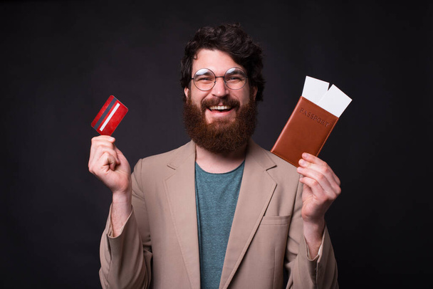 Πορτρέτο του ευτυχισμένη γενειοφόρος hipster άνθρωπος που κατέχουν εισιτήρια διαβατήριο και πιστωτική κάρτα. - Φωτογραφία, εικόνα