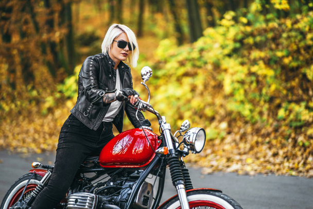 Pretty blonde biker girl in sunglasses with red motorcycle on the road in the forest with colorful blurred background. - Photo, image