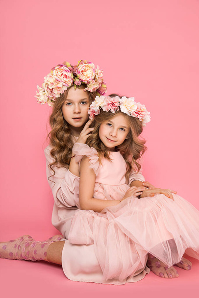 Foto von zwei schönen Mädchen mit Blumenkränzen auf dem Kopf auf rosa Hintergrund, sanfte Umarmungen, Berührungen. Familienbeziehungen zwischen Schwestern. Freundschaft und Liebe. Annäherung. Kinderpsychologie - Foto, Bild